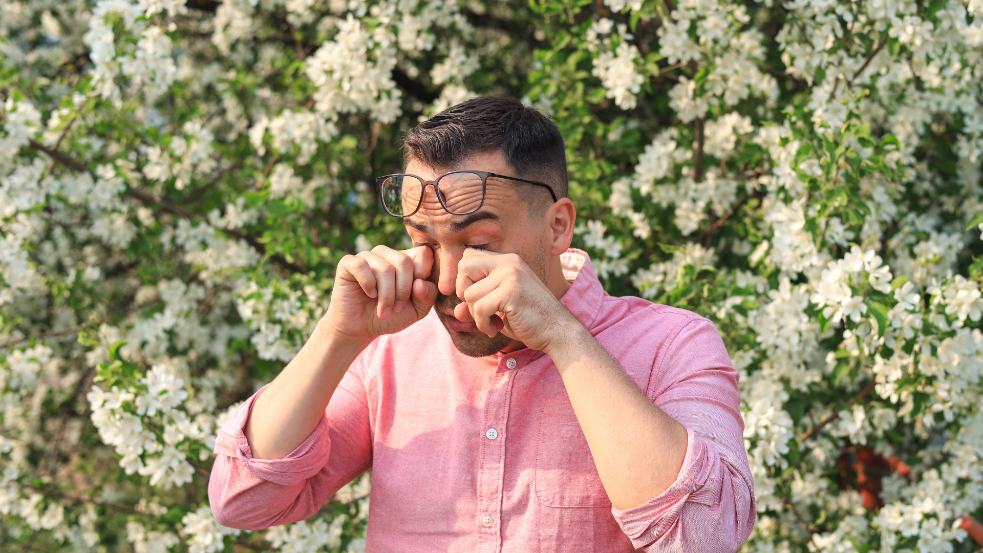 A man standing outside surrounded by flowers rubbing his eyes due to allergies.